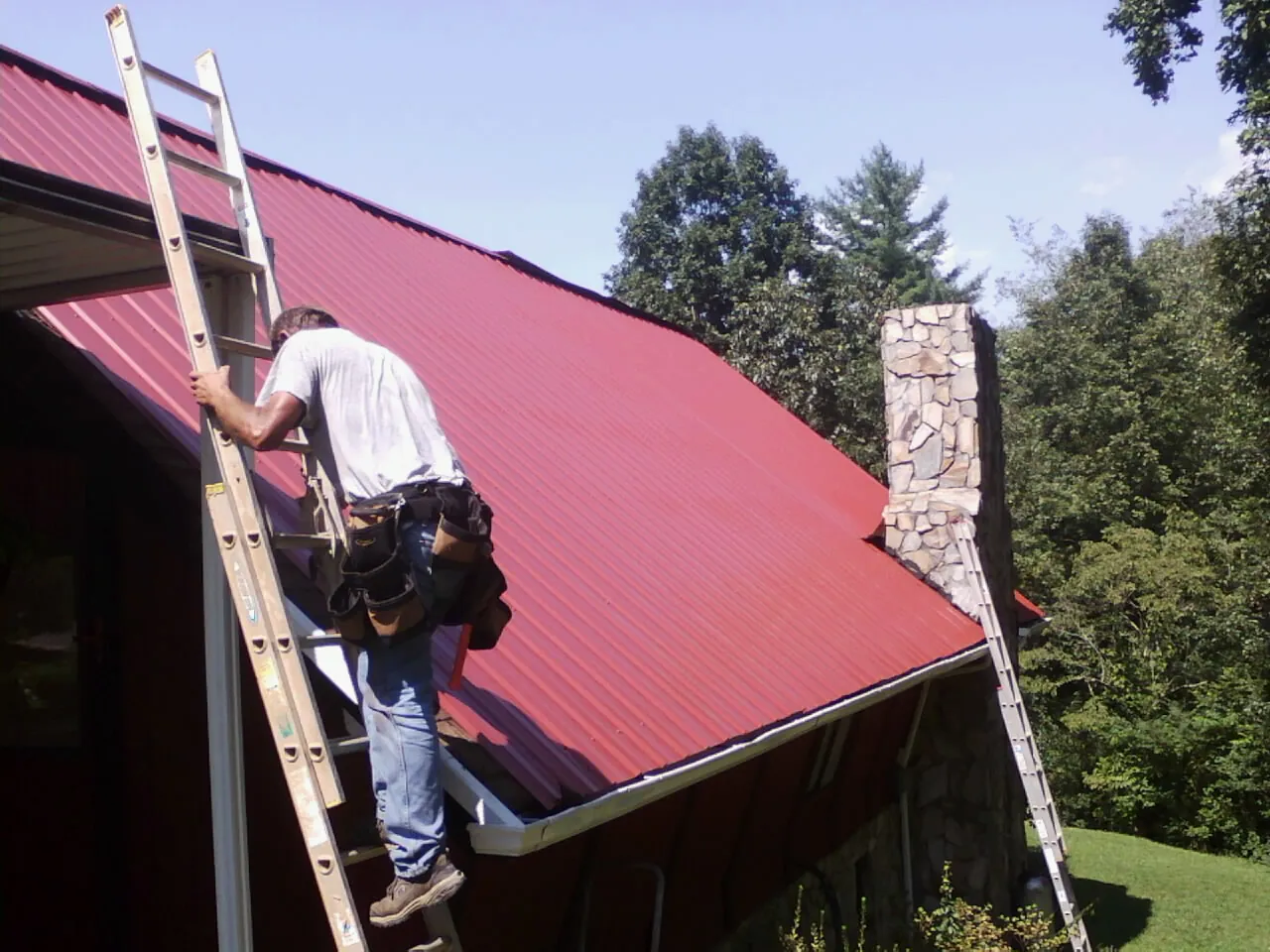 Roofer on a ladder beside a red metal roof and stone chimney — Soffit & Fascia Repair by Roofing Brothers in New Liberty